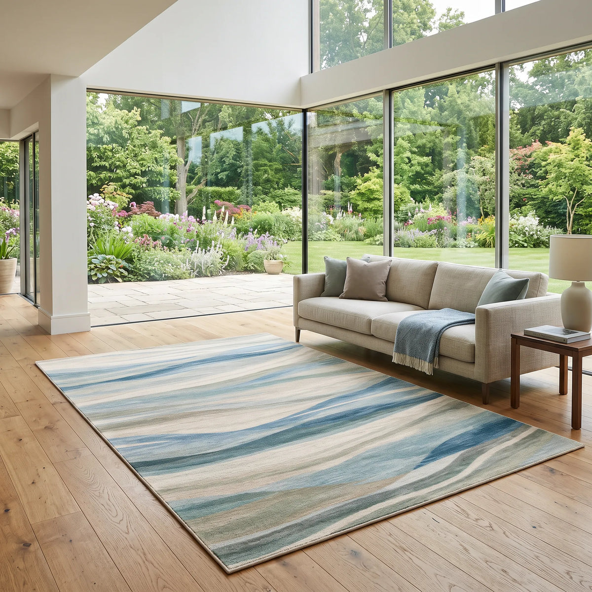 Ein großer rechteckiger Teppich mit einem wellenförmigen abstrakten Muster in Blau-, Beige- und Grautönen, arrangiert in einem modernen Wohnzimmer mit einem hellbeigen Sofa und einem Holzboden. Durch die großen bodentiefen Fenster ist ein üppiger grüner Garten zu sehen.